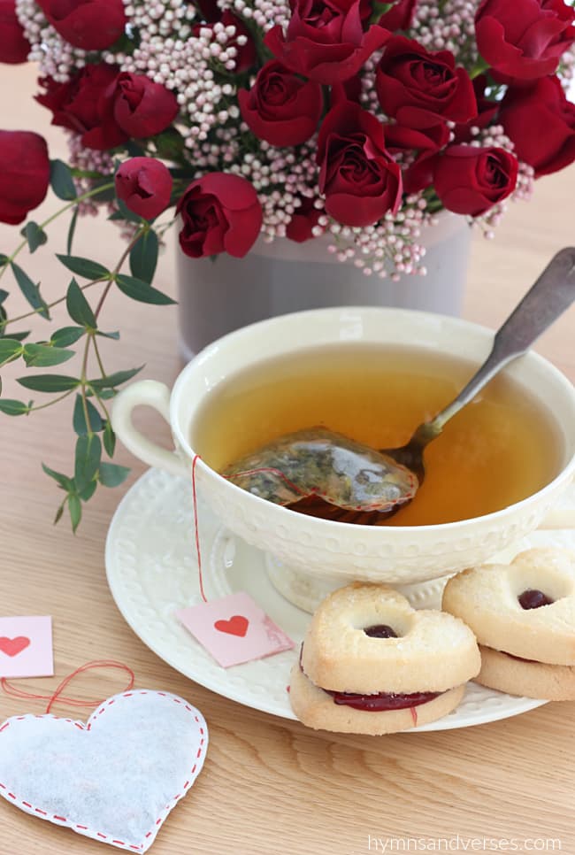 Tea cup with tea and heart shaped tea bag.  Red spray rose bouquet in the background.  Heart shaped cookies on the saucer.
